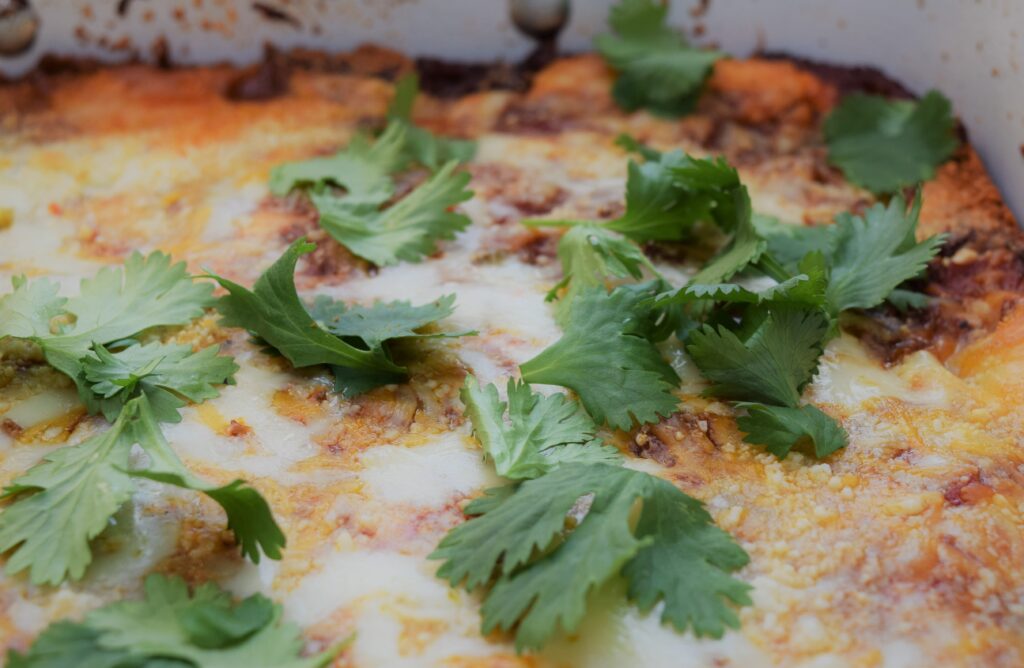 mexican lasagna comfort food spicy hispanic casserole cilantro limes close up in whiye pan, food photography, delicious, decadent, fresh, green, cheesy